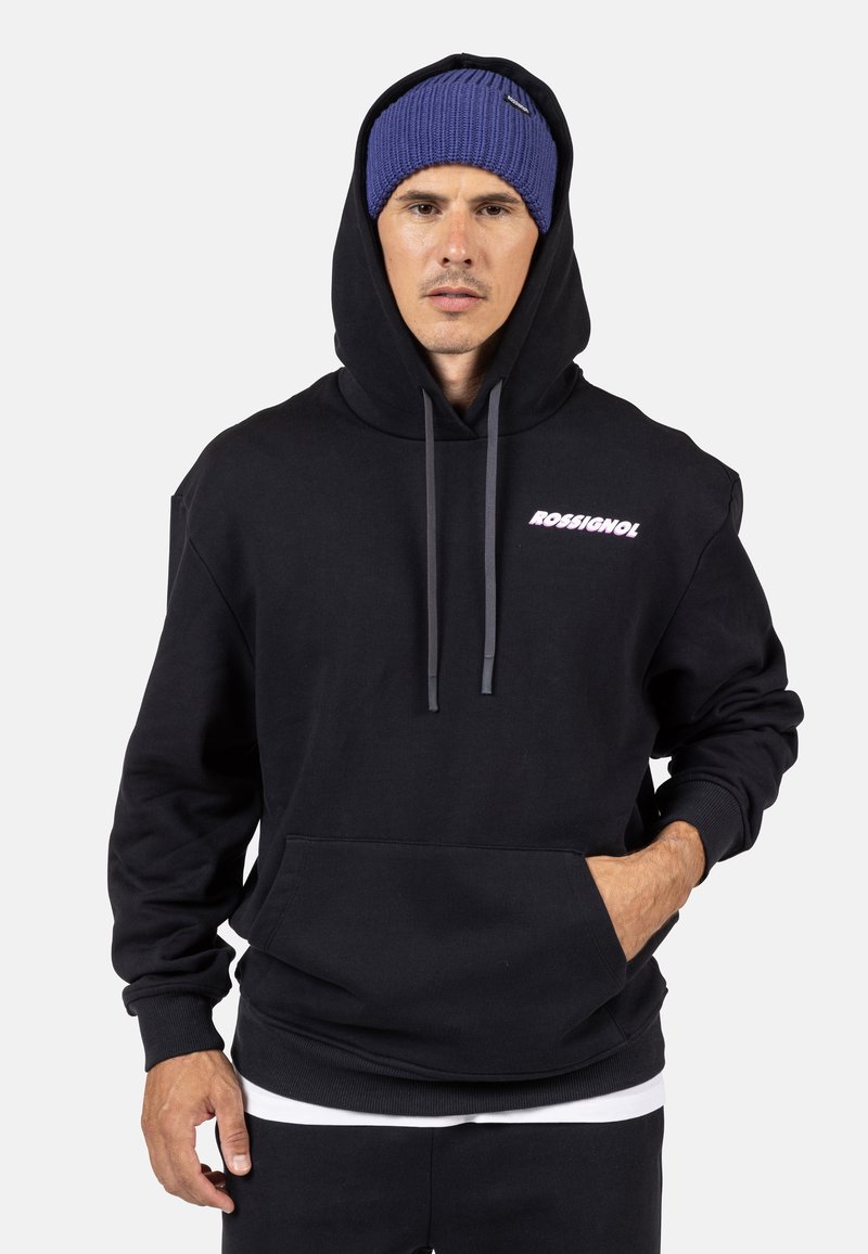 Felpa nera con tasca frontale, cappuccio con cordoncino e logo "ROSSIGNOL" in rosa chiaro. Il modello indossa anche un berretto di maglia blu.