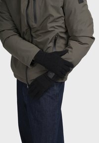 Svarta stickade handskar med texturerad tyg, med en ribbad mudd och en logotypapplikation. Matchas med en grå jacka och denimjeans.