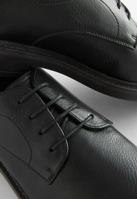 Zapatos de vestir de cuero negro con textura suave, punta afilada y diseño de cordones clásico. Presentan costuras sutiles y un tacón bajo.