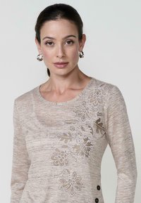 Beige langärmliges Oberteil mit floraler Stickerei in metallischen Tönen, runder Halsauschnitt, horizontaler Struktur und Knopfakzenten an der Seite.