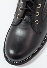 Černé kožené kotníkové boty s kulatou špičkou, které mají černé tkaničky, zlaté očko a ozdobný hřebíkový lem podél podrážky pro zvýraznění detailu.