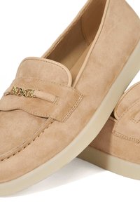 Beige suede slip-on loafers with a gold accent, rounded toe, and a thick cream sole. Smooth texture, stitched detailing on upper.