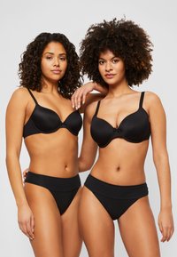 Zwei Modelle tragen abgestimmte schwarze BHs und hochtaillierte Unterhosen. Die BHs verfügen über ein glattes Design mit verstellbaren Trägern und einem kleinen Schleifendetail.
