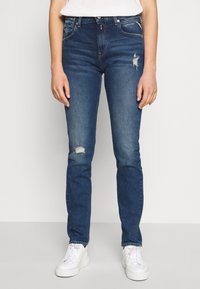 Tmavě modré džíny s úzkým střihem, které mají vybledlé detaily na kolenou a klasický design se pěti kapsami. V kombinaci s bílými teniskami.