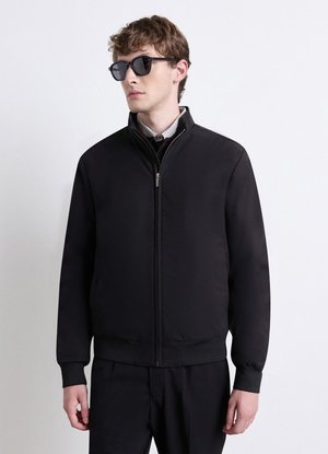 Young man wearing black zip-up jacket, sunglasses, and dark trousers, standing against a plain light gray background.