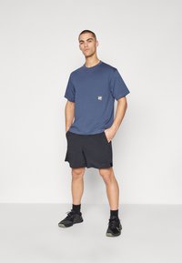 Tricou bleumarin cu mâneci scurte și un mic patch cu logo, purtat cu pantaloni scurți negri și pantofi sport negri. Design simplu, casual.