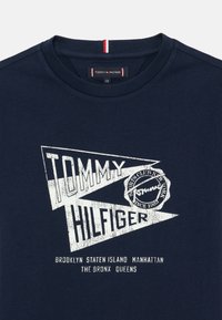 Námořnická modrá tričko s bílým grafickým textem "TOMMY HILFIGER" a designem razítka. Kulatý výstřih s pruhovaným červeným a bílým akcentem.