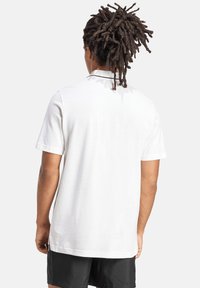 Weißes Polohemd mit kurzen Ärmeln, Kragen und schwarzen Details, aus weichem Stoff. Zu sehen ist eine Person, die es trägt, von hinten betrachtet.