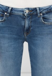 Plave traperice s srednjim strukom, pet džepova i kontrastnim šavovima. Imaju kopčanje na dugme i blago izbjeljivanje tkanine.