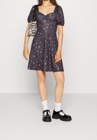 Černé květinové šaty s nabíranými rukávy, čtvercovým výstřihem a rozprašující sukní. Doplňky zahrnují pruhovanou kabelku a černé chunky boty s bílými ponožkami.