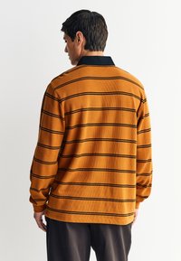 Košulja dugih rukava s ovratnikom u boji opečene naranče s crnim horizontalnim prugama. Ima teksturiranu tkaninu i spuštena ramena.