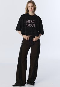 Maglietta nera corta con texture a volant e scritta "MERCI AMOUR" in lettere rosa, abbinata a pantaloni larghi marrone scuro con strisce laterali.