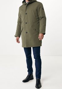 Olivgrön parka med huva, med knappstängning, två framfickor och svarta accenter. Buren över marinblå jeans och svarta stövlar.