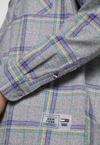 Manica di camicia a quadri grigi con linee blu, gialle e verdi, polsino abbottonato e etichetta Tommy Jeans vicino all'anca.