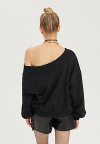 Musta ylisuuri collegepaita olkapäät paljastavalla designilla, resorit hihansuissa ja helmassa. Yhdistetty kiiltäviin mustiin shortseihin ja kalaverkko-sukkahousuihin.