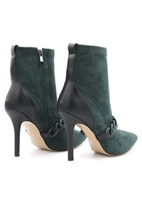 Stivaletti alla caviglia in suede verde con un dettaglio in pelle nera, punta affusolata e una catena decorativa, montati su alti tacchi a spillo.