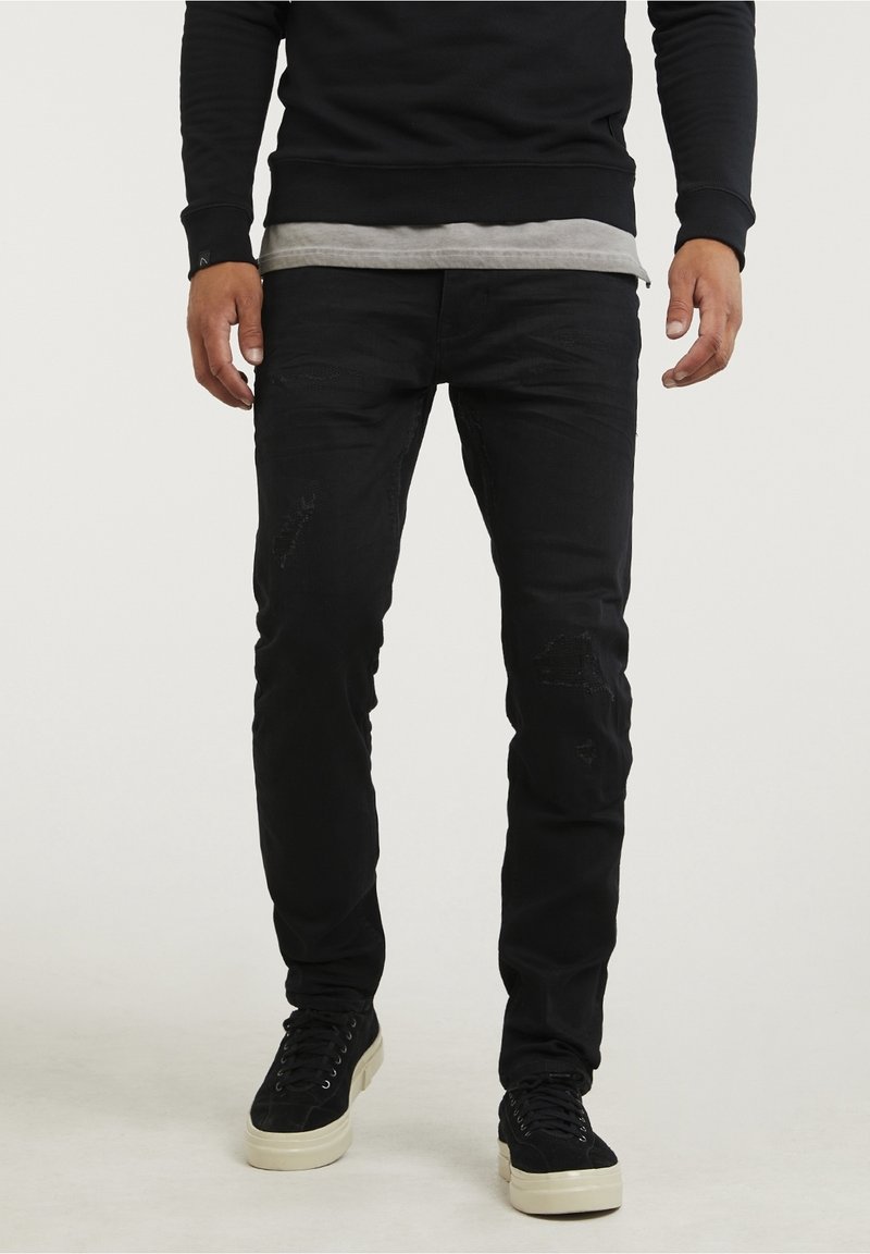 Homme portant un jean noir slim avec légère usure, un sweat-shirt noir et des baskets noires à semelles blanches, debout sur un fond clair uni.