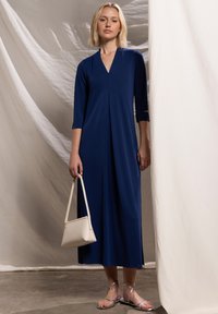 Marineblauwe jurk met een V-hals en driekwartmouwen, voorzien van plooien en een vloeiende silhouet. Gecombineerd met een kleine witte handtas.