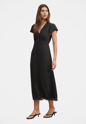 Femme portant une robe midi noire à petits pois blancs avec des boutons sur le devant, associée à des sandales noires à talons.