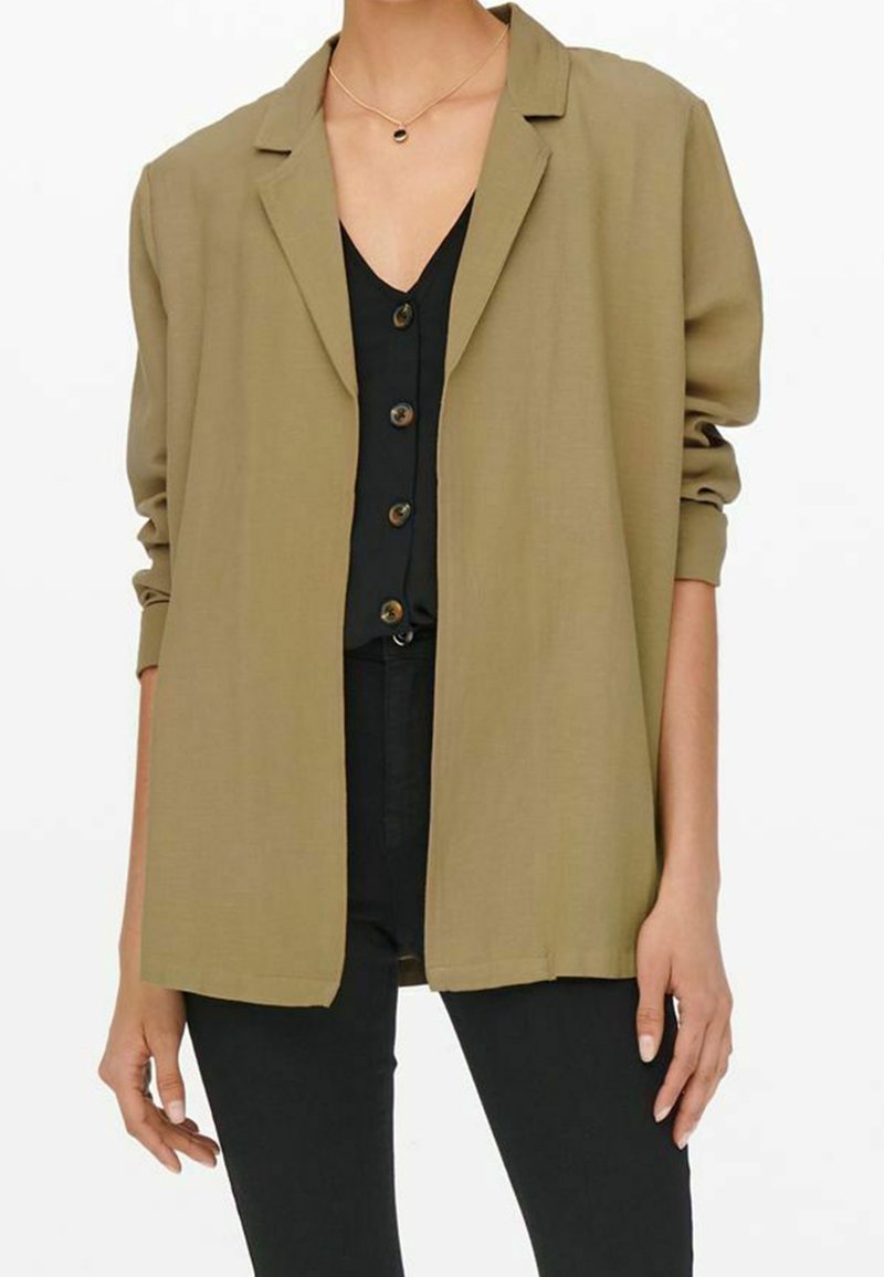 Donna che indossa un blazer oversize marrone chiaro sopra una maglietta nera con bottoni e pantaloni neri, accessorita con una semplice collana d'oro.