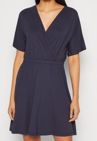 Granatowa sukienka kopertowa z krótkimi rękawami, dekoltem w serek, elastyczną talią i zwiewnym dołem. Wykonana z miękkiego materiału o gładkiej strukturze.