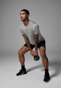Szara koszulka sportowa z krótkim rękawem, czarne szorty i czarne buty sportowe. Trzyma czarny kettlebell, widoczne tatuażowane ramiona.