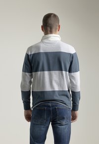 Camisa polo de manga larga para hombre con rayas horizontales en gris oscuro y claro, un cuello blanco y puños acanalados. Se lleva con jeans azules.