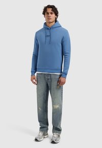 Blauwe katoenen hoodie met een voorzak, een trekkoordcapuchon en geborduurd logo. Gepaard met licht versleten denim jeans en sneakers.