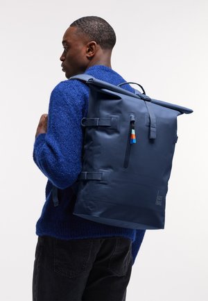 Young man in blue sweater carrying large navy roll-top backpack with front zipper and colored zipper pull tabs.
