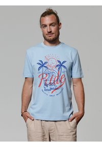 Lichtblauw T-shirt met korte mouwen van katoen, met een grafisch ontwerp van een zon, palmboompjes en de tekst "Ride" in rood en blauw.