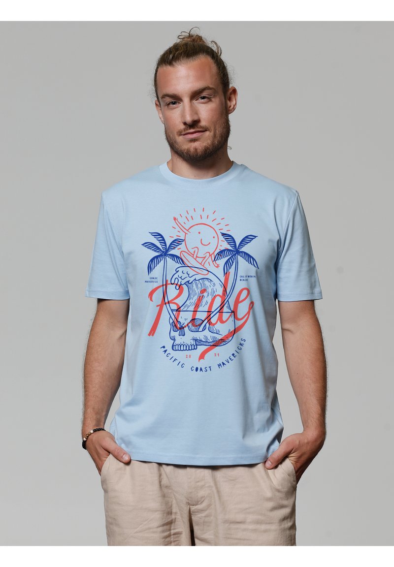 Lichtblauw T-shirt met korte mouwen van katoen, met een grafisch ontwerp van een zon, palmboompjes en de tekst "Ride" in rood en blauw.