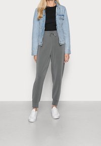 Kvinna som bär en ljus denimjacka, svart topp, grå joggers med snörning och vita sneakers, stående mot en enkel ljus bakgrund.