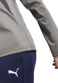 Maglia grigia a maniche lunghe con finitura testurizzata e pannelli in rete. Presenta l'etichetta "DRYCELL" sull'orlo, abbinata a pantaloni blu navy e un logo bianco.