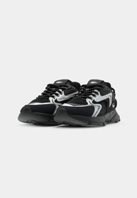 Zapatillas deportivas negras con parte superior de malla, acentos plateados, superposiciones texturizadas, suela de goma negra gruesa y sistema de cordones.