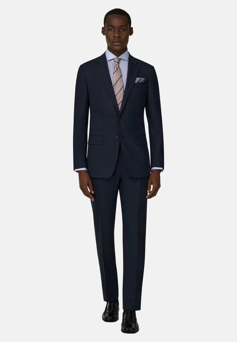 Homme debout, portant un costume bleu marine foncé avec une cravate beige rayée, une chemise bleu clair, un mouchoir de poche à motifs, et des chaussures habillées noires sur fond blanc.
