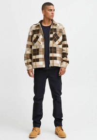 Brun og beige rutete flanellskjorte med to brystlommer, brukt over en svart t-skjorte, sammen med mørke jeans og brune støvler.