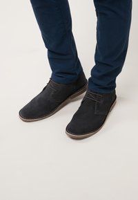 Botas de ante oscuras hasta el tobillo con punta redonda y detalles de cordones de cuero, combinadas con pantalones slim-fit de color azul marino.
