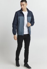 Tengerészkék és világoskék szélálló dzseki, cipzárral az elején, rugalmas mandzsettával és aljjal. Fehér póló és fekete nadrág felett viselve. Fekete sneaker cipő.