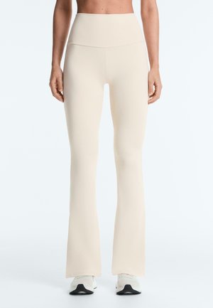 Ženska, ki nosi visokodežne, bež razširjene pajkice in bele športne copate, stoji ob belem ozadju.