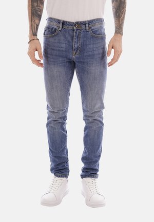 Uomo che indossa jeans slim fit in denim blu, sneakers bianche e una camicia bianca, con tatuaggi visibili sugli avambracci.