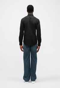 Čierna košeľa s dlhým rukávom a rovnými strihom, nosená s voľnými modrými džínsami. Biele tenisky dotvárajú outfit.