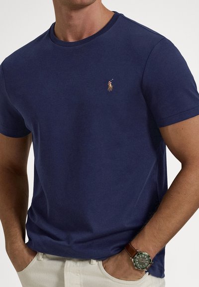 T-shirt in cotone blu navy con scollo rotondo, maniche corte e piccolo logo ricamato sul petto. Indossata con pantaloni chiari.