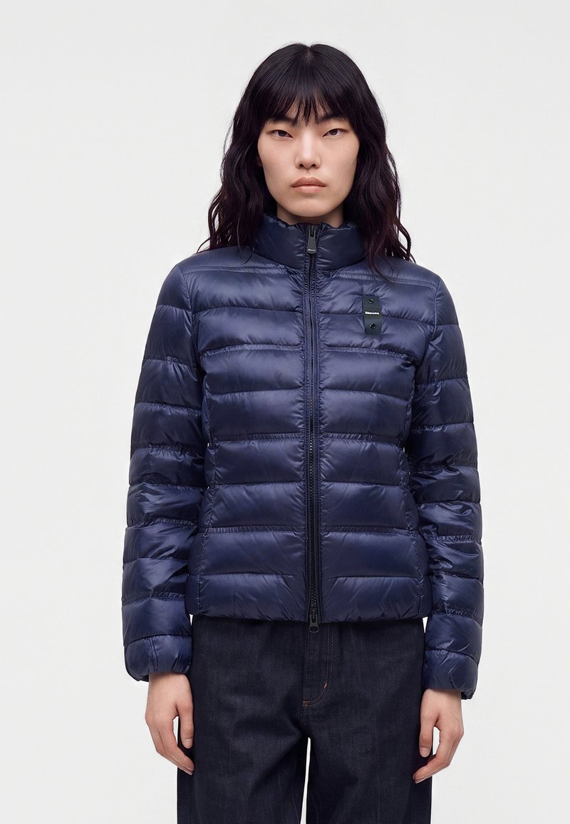 Femme aux longs cheveux foncés porte une veste matelassée bleu marine zippée, debout devant un fond blanc uni.