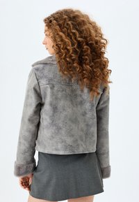 Graue Kunstlederjacke mit einem weichen Fellkragen und -manschetten. Verfügt über eine strukturierte Passform und eine strukturierte Oberfläche. Modell von hinten gezeigt.