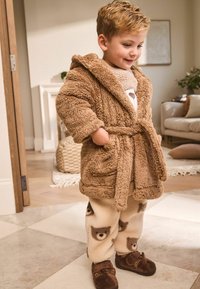 Junges Kind lächelt drinnen, trägt einen flauschigen braunen Kapuzenbademantel, Fleecehosen mit Bärenmuster und braune Hausschuhe mit Bärengesichtern.