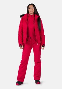Rote isolierte Jacke mit Kapuze und Reißverschluss, kombiniert mit roten Skihosen. Beide haben eine glatte Textur und praktische Taschen. Schwarze Akzente an den Ärmeln.