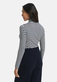 Vrouw met lang donker haar, gekleed in een zwart-wit gestreepte coltrui en donkere high-waisted broeken, zijwaarts gekeerd tegen een witte achtergrond.