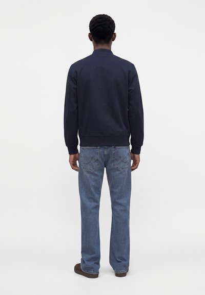 Homme de dos portant une veste bleu marine foncé, un jean bleu clair et des chaussures marron foncé sur un fond blanc uni.