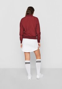 Burgunderfarbene Bomberjacke mit gerippten Bündchen und Saum, kombiniert mit einem weißen Kleid. Das Modell trägt weiße knielange Socken mit schwarzen Streifen und Sneakers.