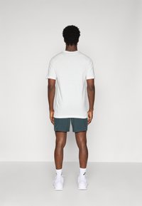 Camiseta de algodón blanca con mangas cortas, combinada con pantalones cortos de color teal. El modelo se encuentra de pie frente a un fondo sencillo, mostrando la vista desde atrás.